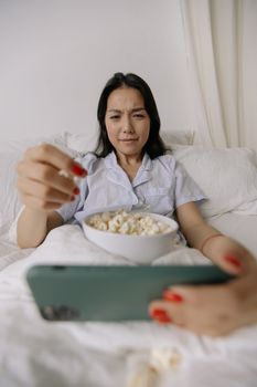 Asian woman in bed with popcorn watching on smartphone, enjoying leisure time.
