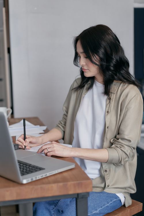 Free A woman sitting at a desk writing in a notebook next to her laptop indoors. Stock Photo