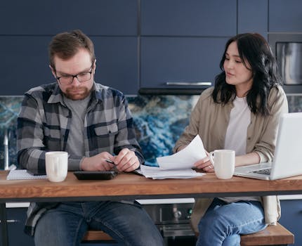 A couple discusses bills and finances using a laptop and calculator in their modern kitchen.