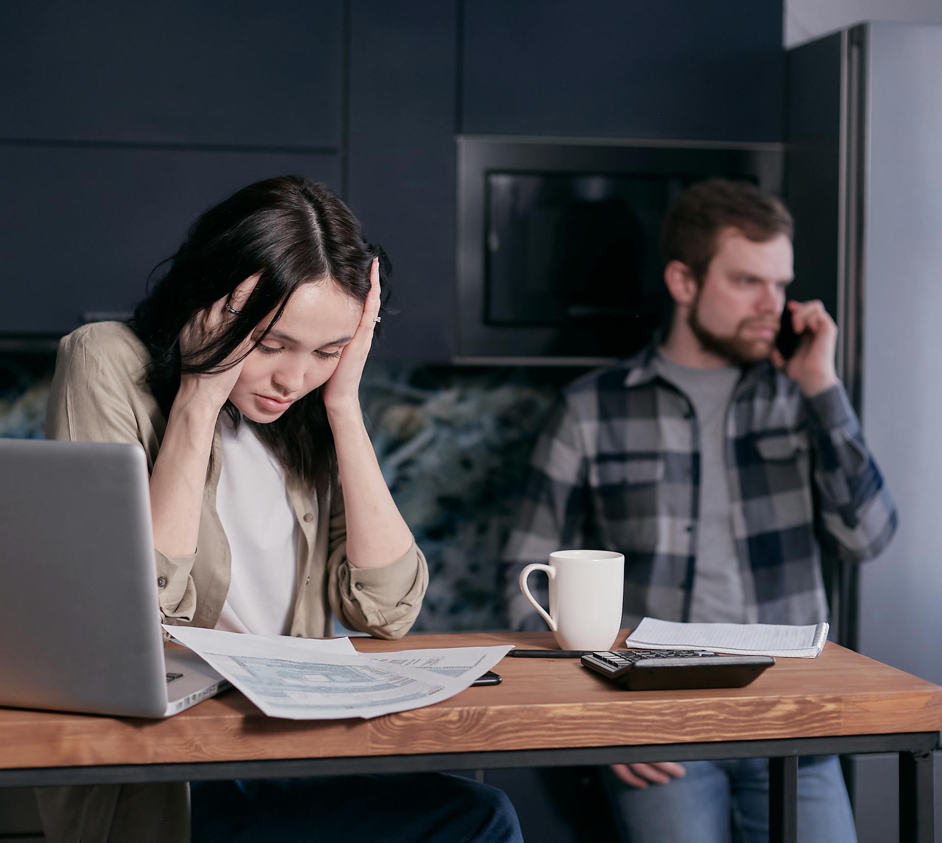 A stressed woman holds her head while working at a desk with documents and a laptop.