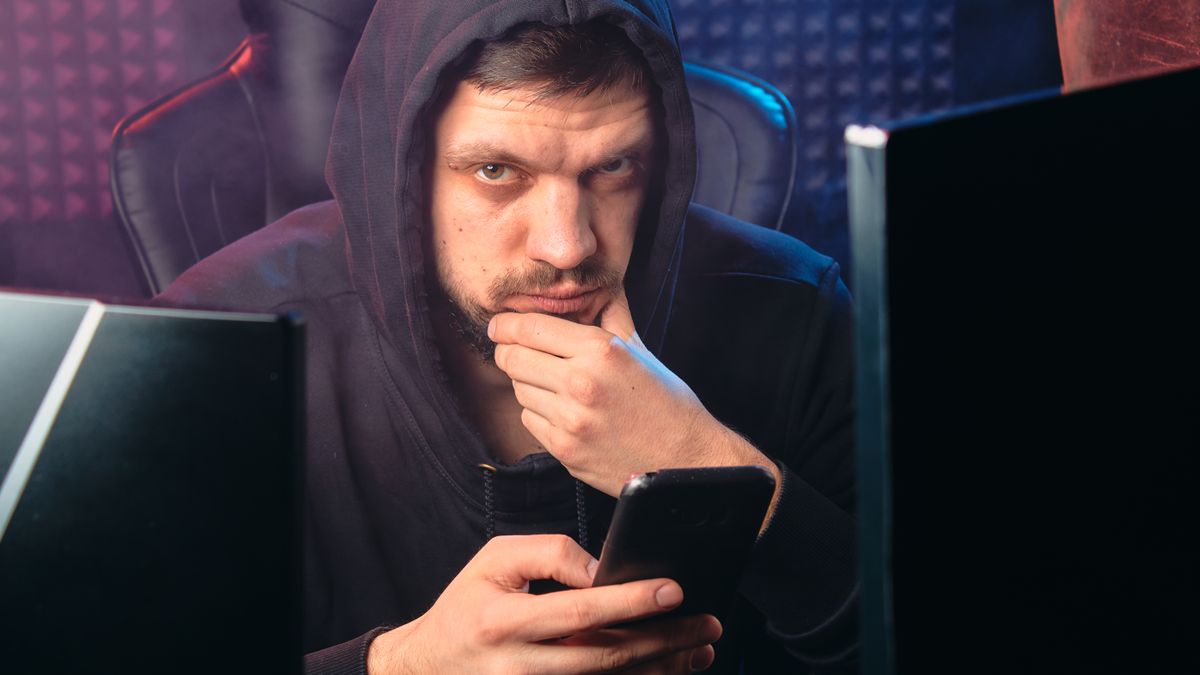 Homem de moletom escuro olhando preocupado para o celular diante de telas digitais, representando uma tentativa de golpe de phishing bancário
