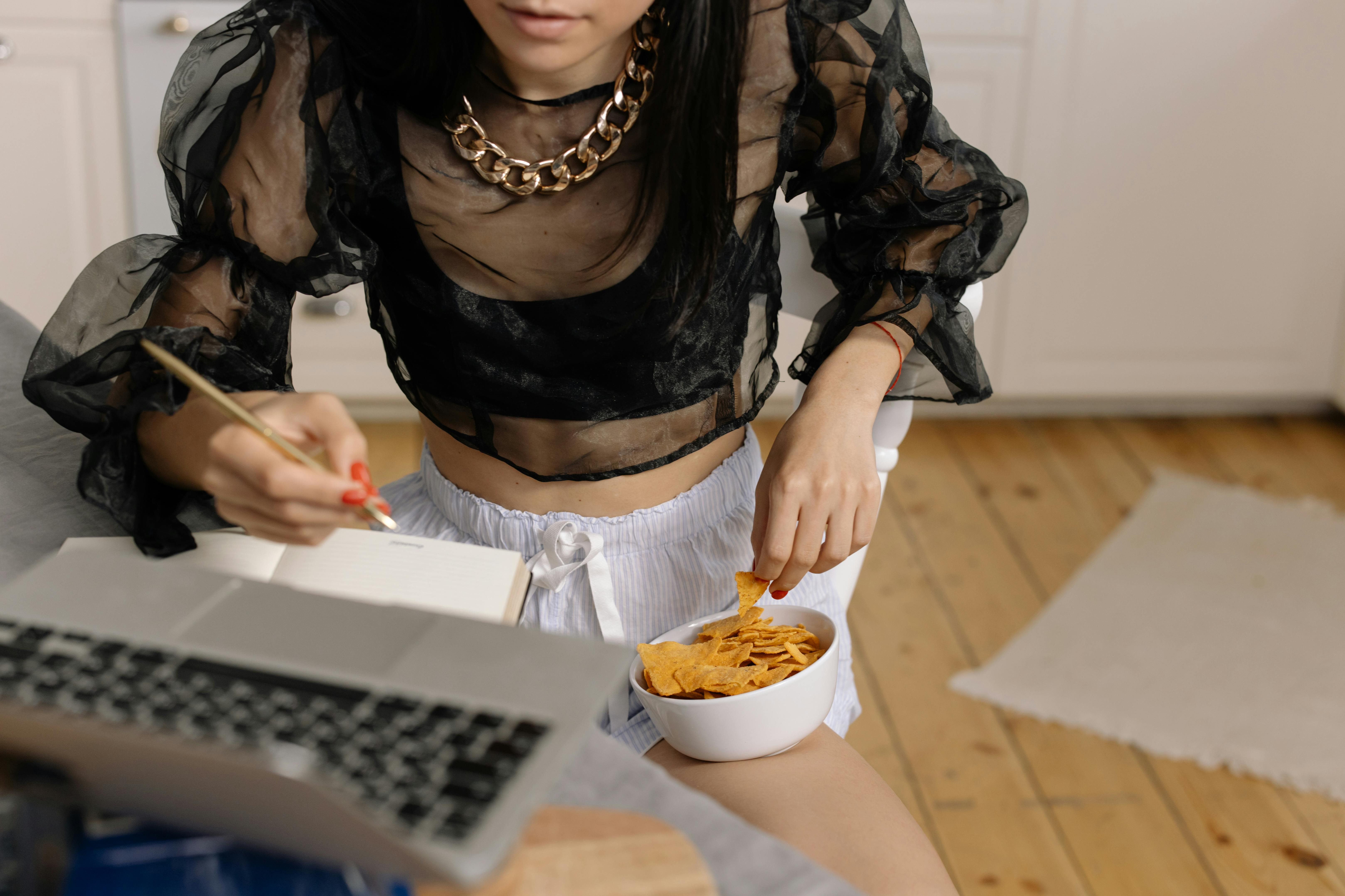 Young woman writing notes while working remotely, enjoying a snack at home.