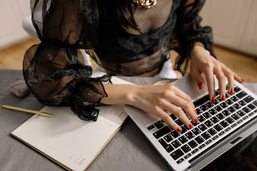 Close-up of a woman working on a laptop with red nails, notebook, and pen.