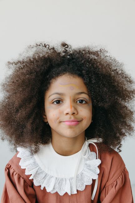 Charming portrait of a young girl with curly hair and face paint, wearing a brown dress with lace detail.