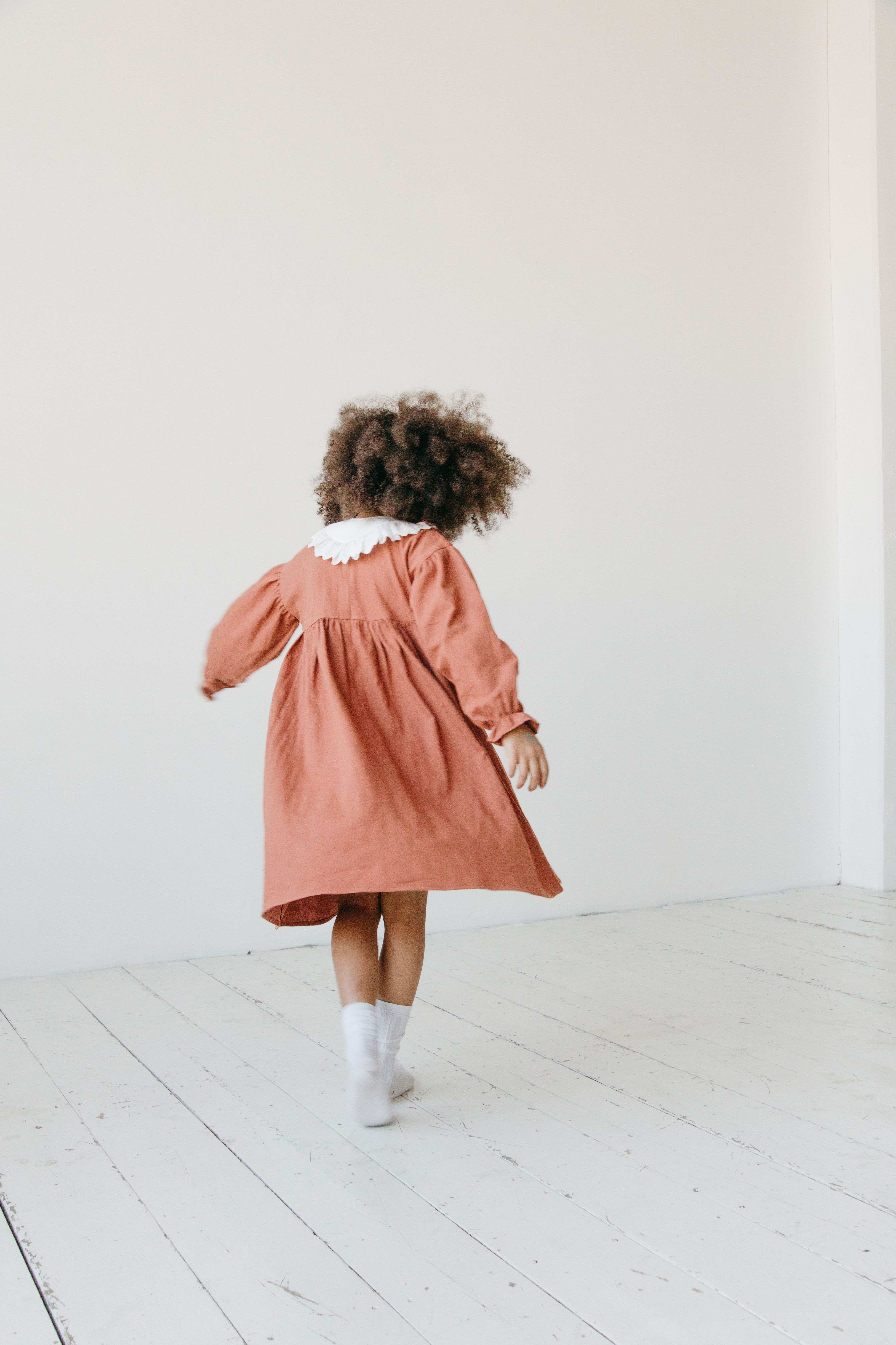 A young girl with curly hair wearing a brown dress and walking indoors. Bright and playful scene.