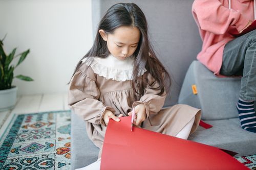 Free Young girl focused on cutting red paper indoors, showcasing creativity and concentration. Stock Photo