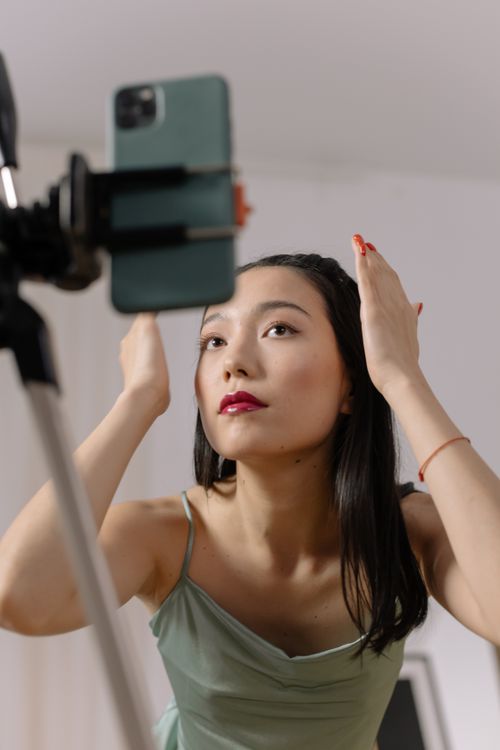 Free Asian woman adjusts hair and makeup in front of smartphone for live online content creation. Stock Photo