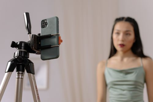 Free A woman stands in front of a smartphone set up on a tripod, engaged in vlogging content indoors. Stock Photo