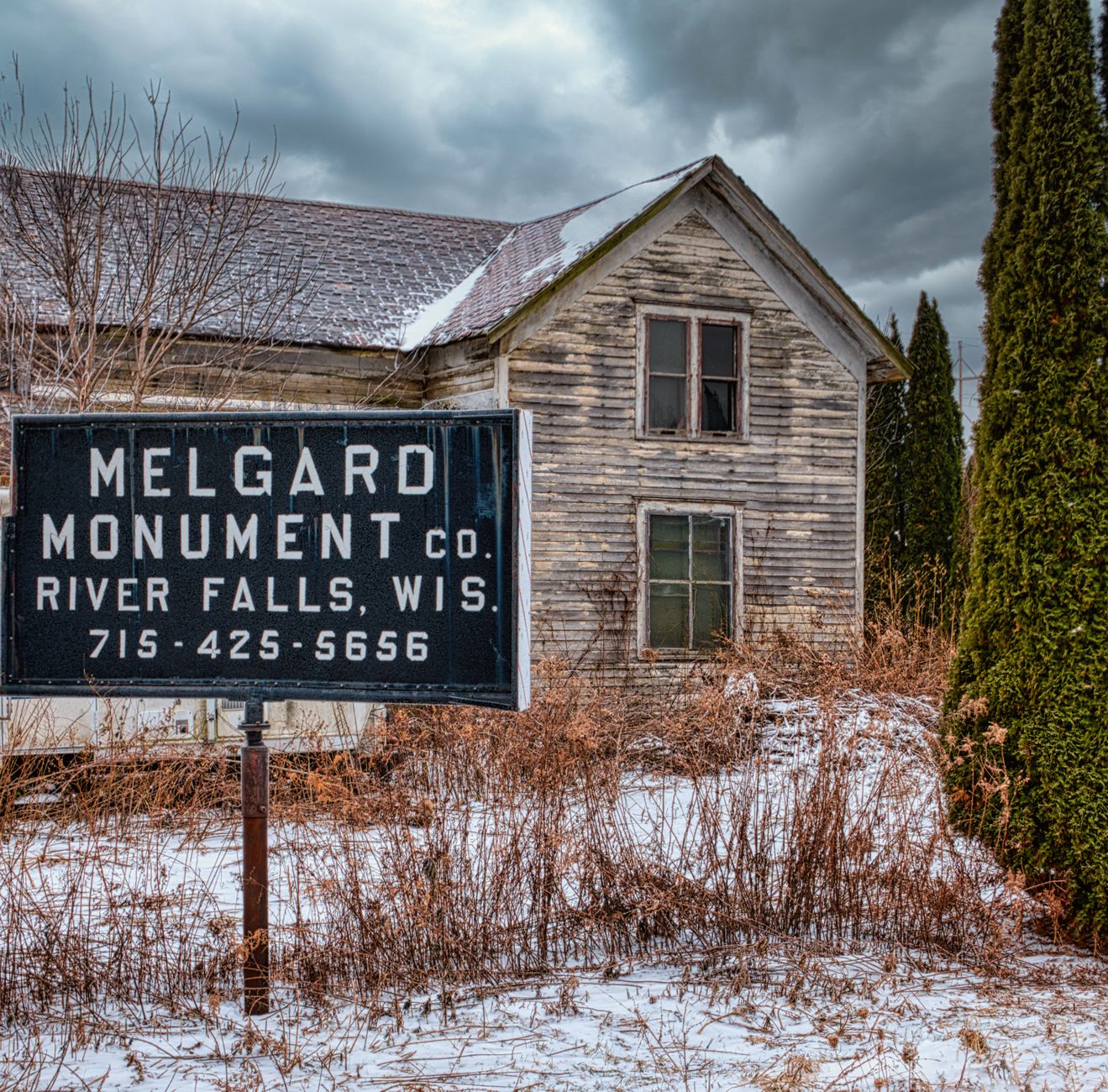 Abandoned wooden house with aged exterior and Melgard Monument Co. signage in snowy landscape.