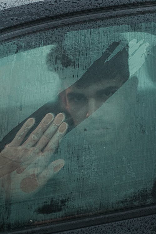 Free Through glass of serious ethnic man looking at camera through wet window with trace of hand while sitting in automobile in rainy weather Stock Photo