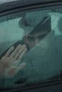 Through glass of serious ethnic man looking at camera through wet window with trace of hand while sitting in automobile in rainy weather