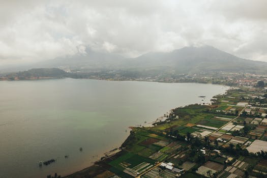 Stunning aerial view of a lake surrounded by lush green fields and mountains under a cloudy sky.