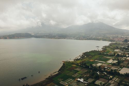 Free Stunning aerial view of a lake surrounded by lush green fields and mountains under a cloudy sky. Stock Photo