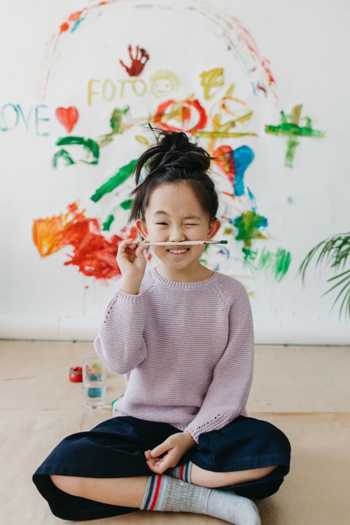 Free A joyful child winks while holding a brush, surrounded by colorful wall art indoors. Stock Photo