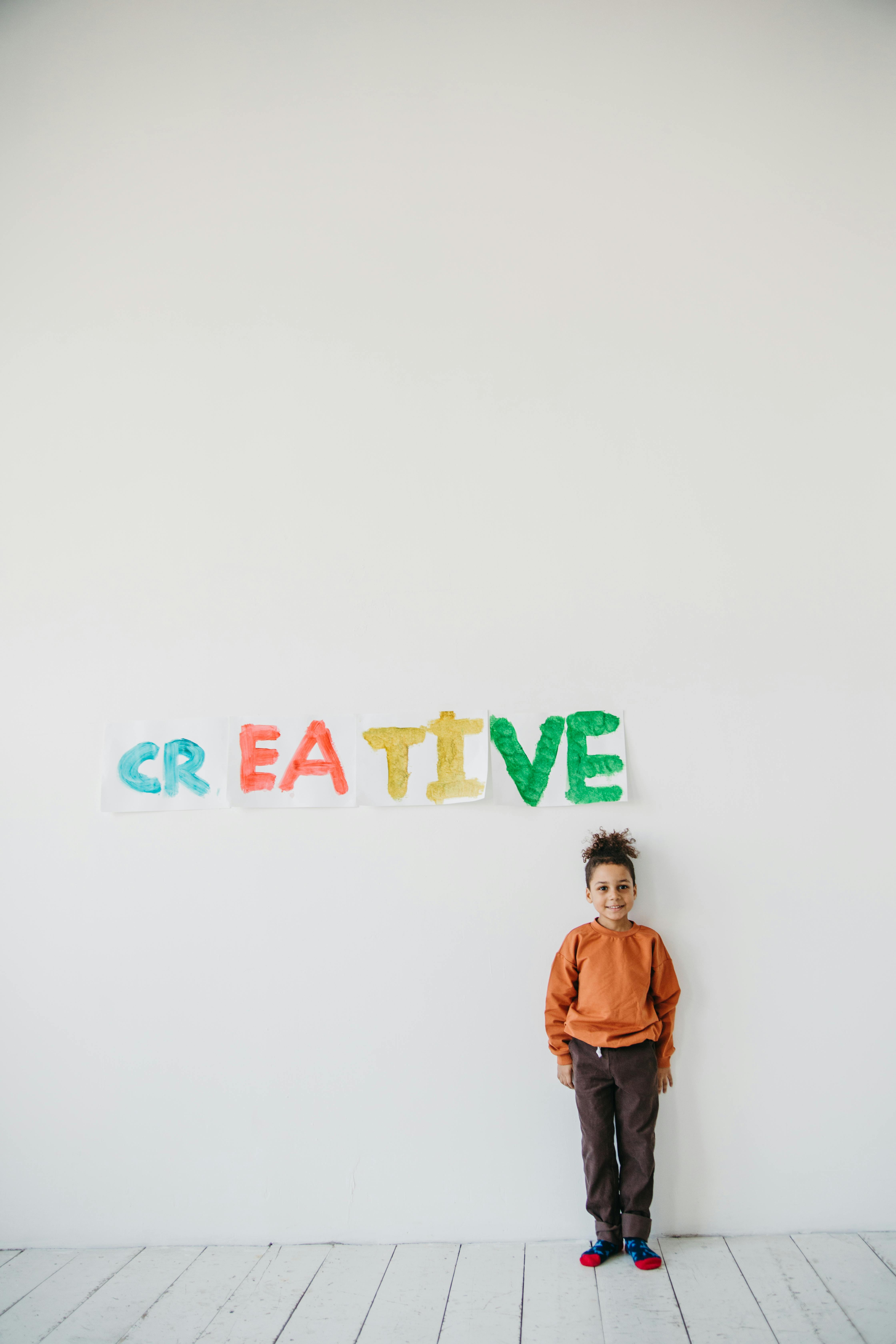 Young boy stands by a colorful "creative" sign on a white wall, embodying youthful imagination.