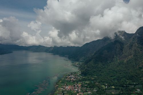 Free A breathtaking aerial view showcasing lush green mountains, a serene lake, and a small village under a cloudy sky. Stock Photo