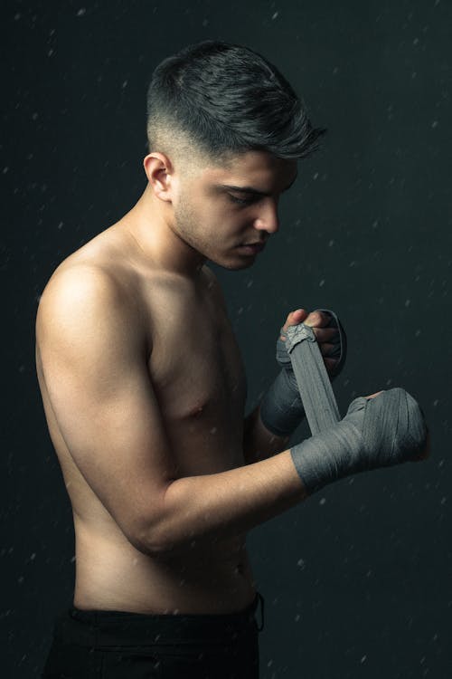 Side view of muscular young male fighter with naked torso wrapping hands with protective elastic band before workout against black background