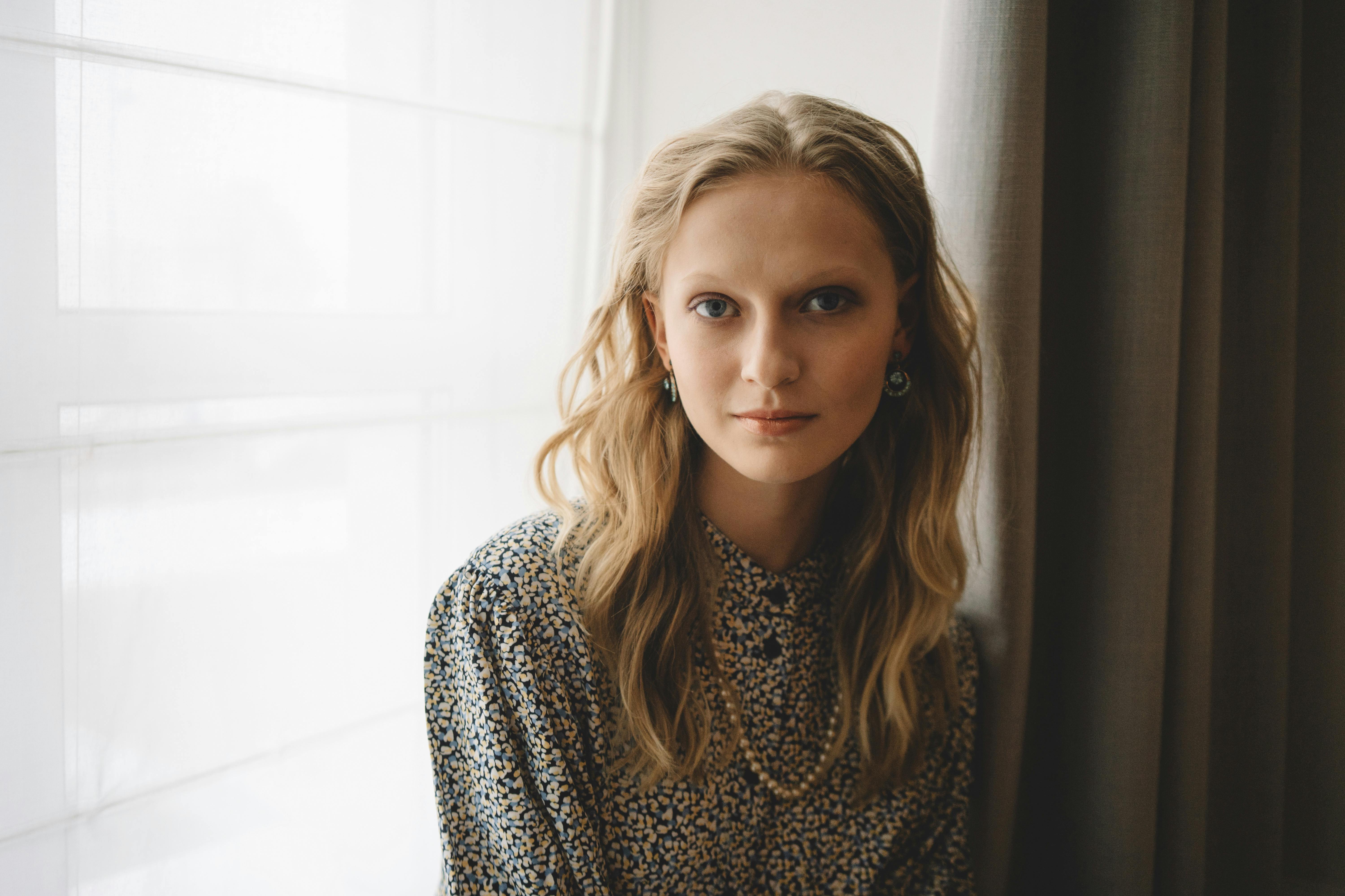 Portrait of a fashionable woman with blonde hair near a window indoors.