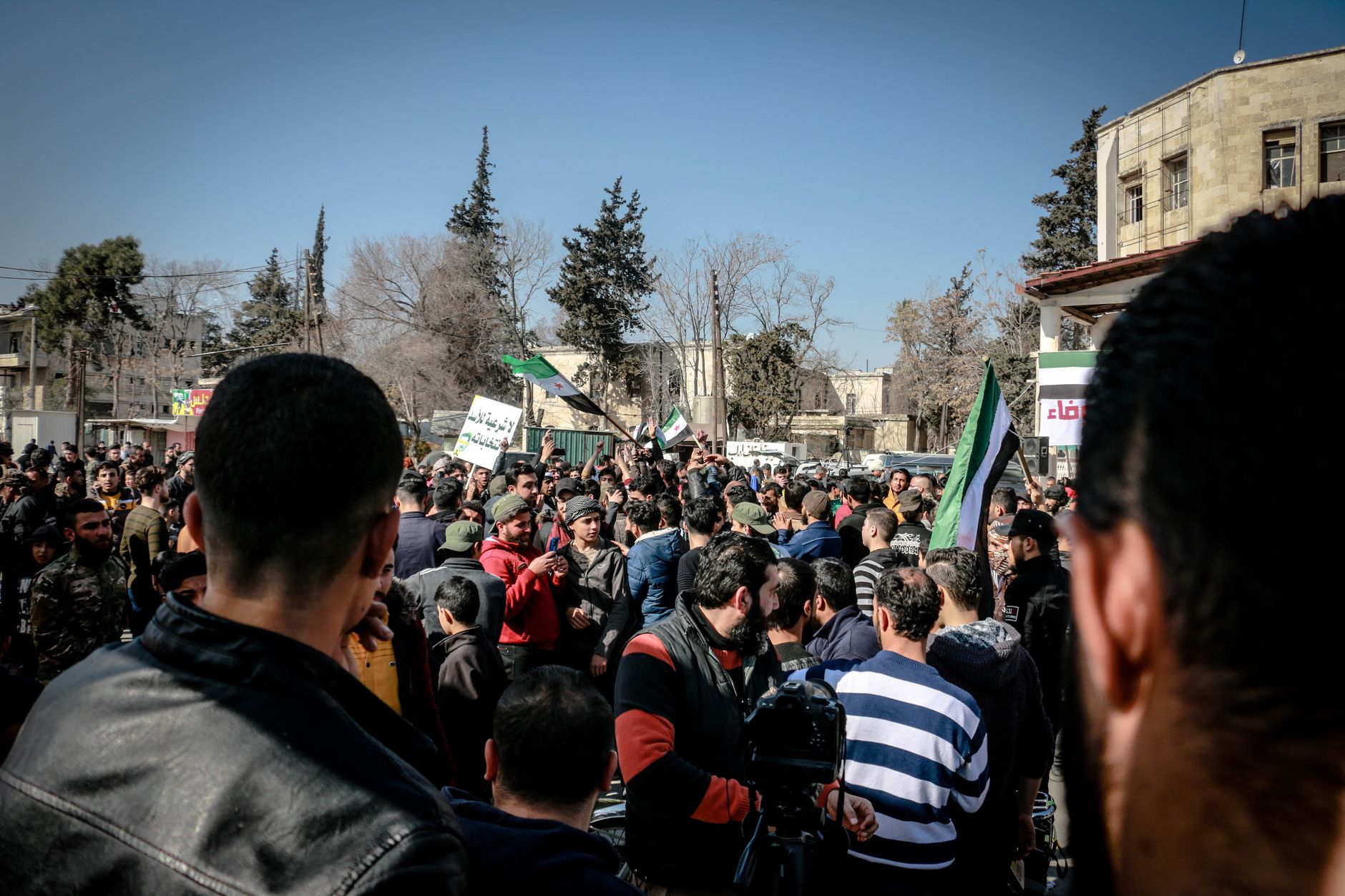 A large crowd gathers outdoors for a protest in Idlib, Syria under a clear sky.