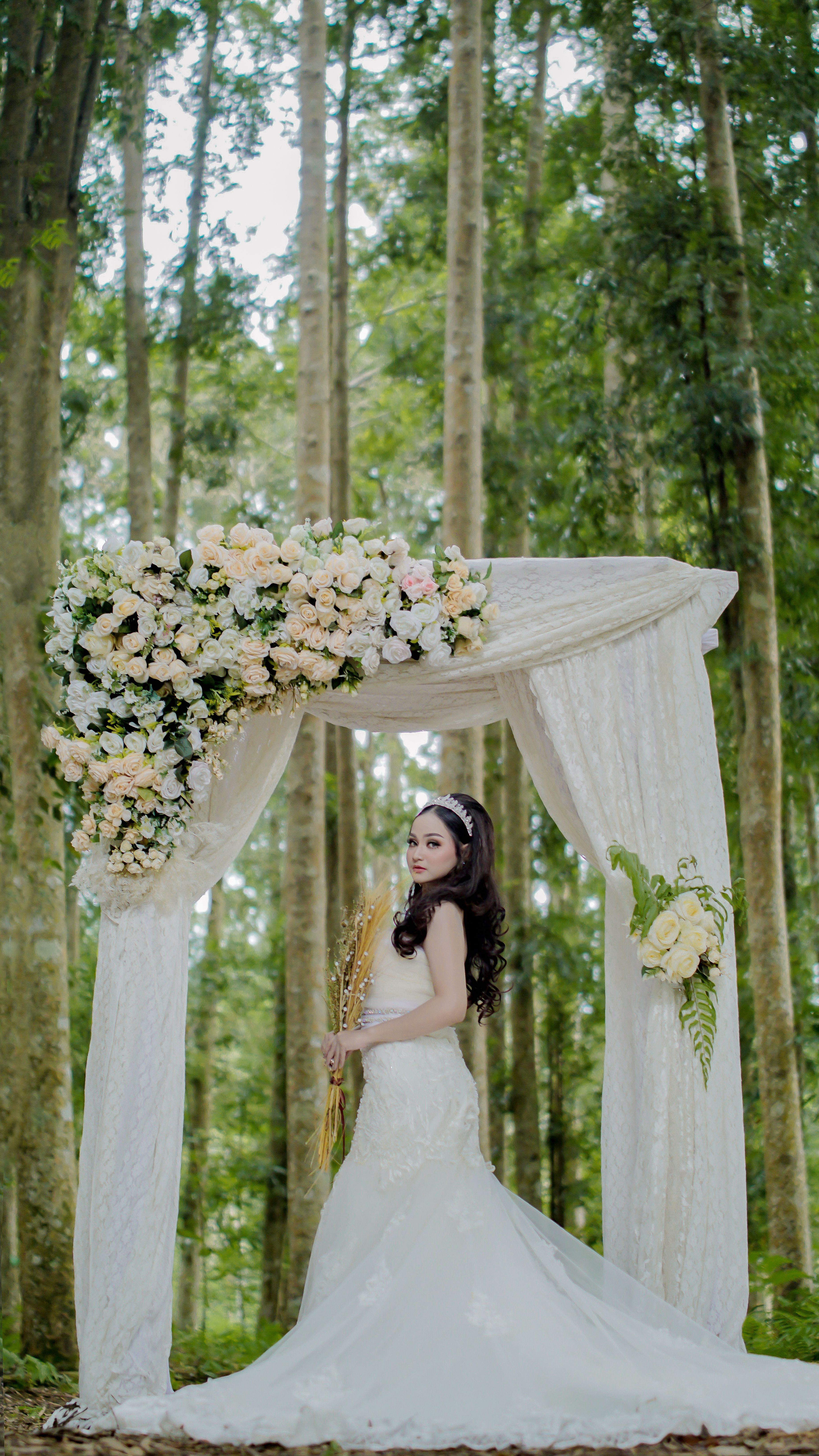 A bride poses under a floral arch in a serene forest setting, capturing a beautiful wedding moment.