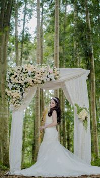 A bride poses under a floral arch in a serene forest setting, capturing a beautiful wedding moment.