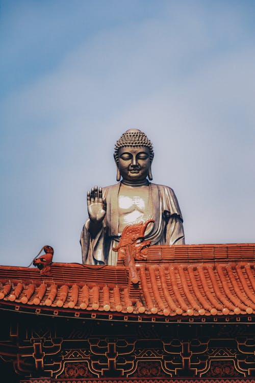 From below of aged stone Buddha statue against ornamental roof of traditional oriental temple against cloudless blue sky