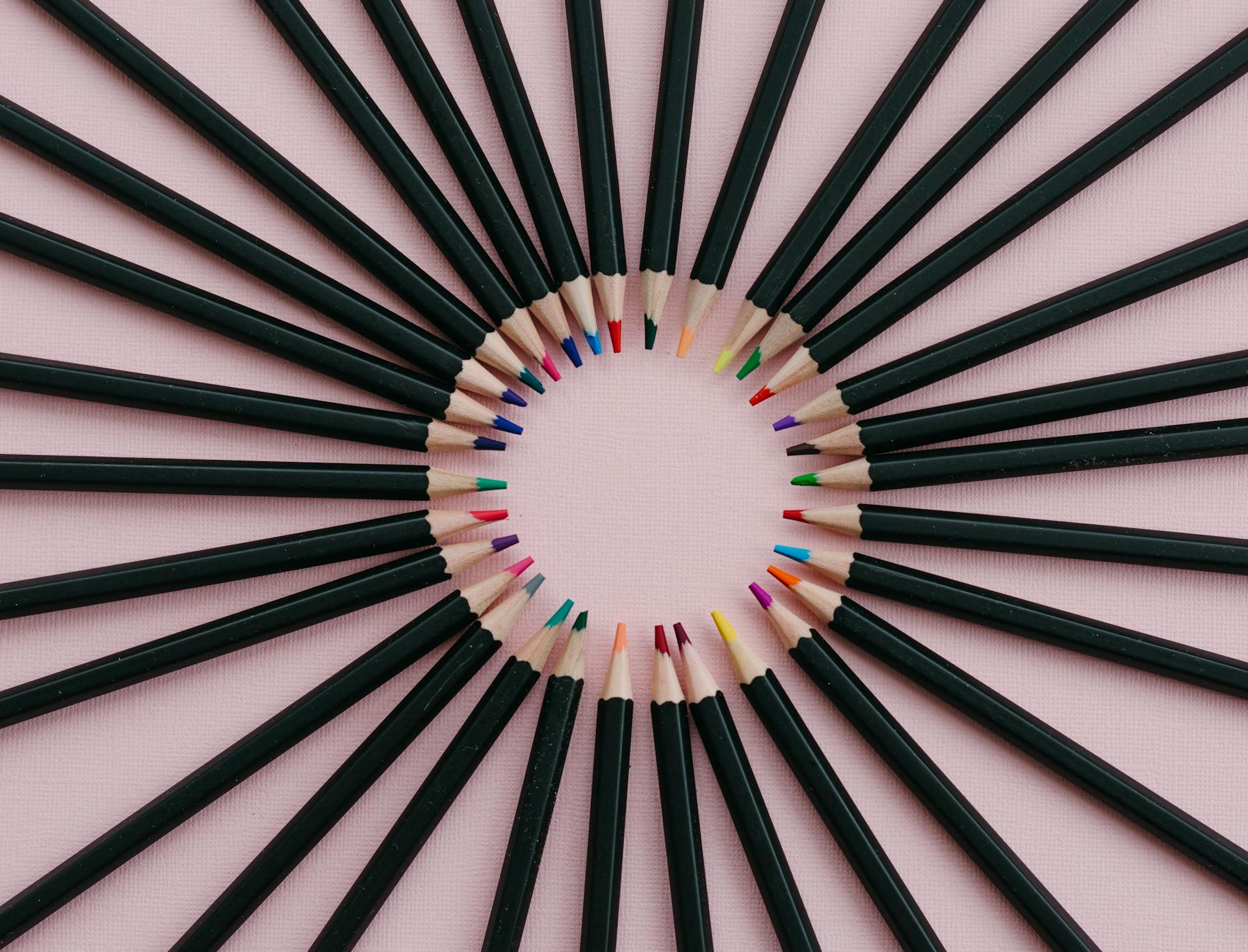 Artistic arrangement of colored pencils fanned out on a pink surface, creating a radial pattern.