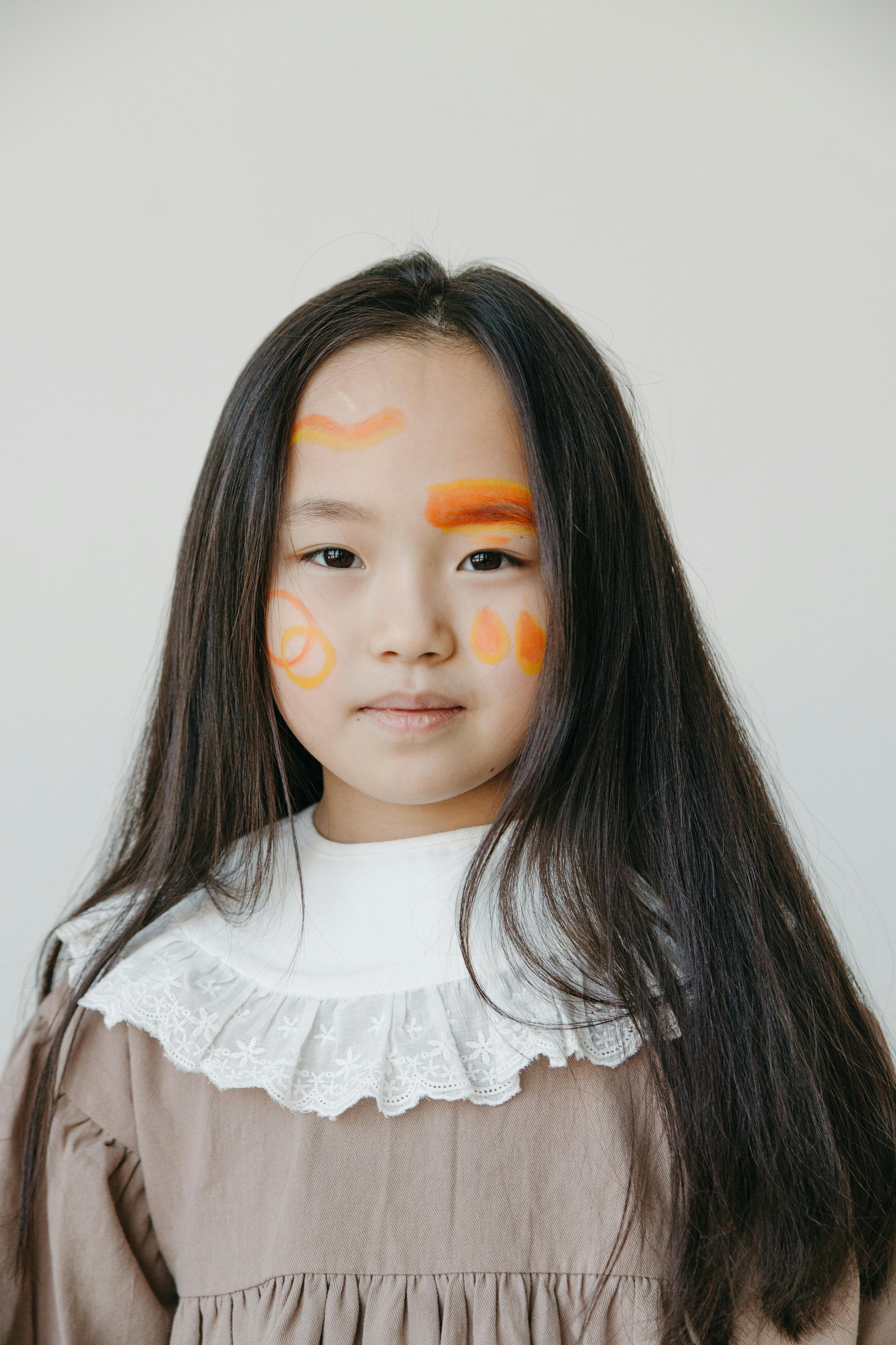 Portrait of a young girl with artistic face paint and long hair, wearing a brown dress with lace collar.