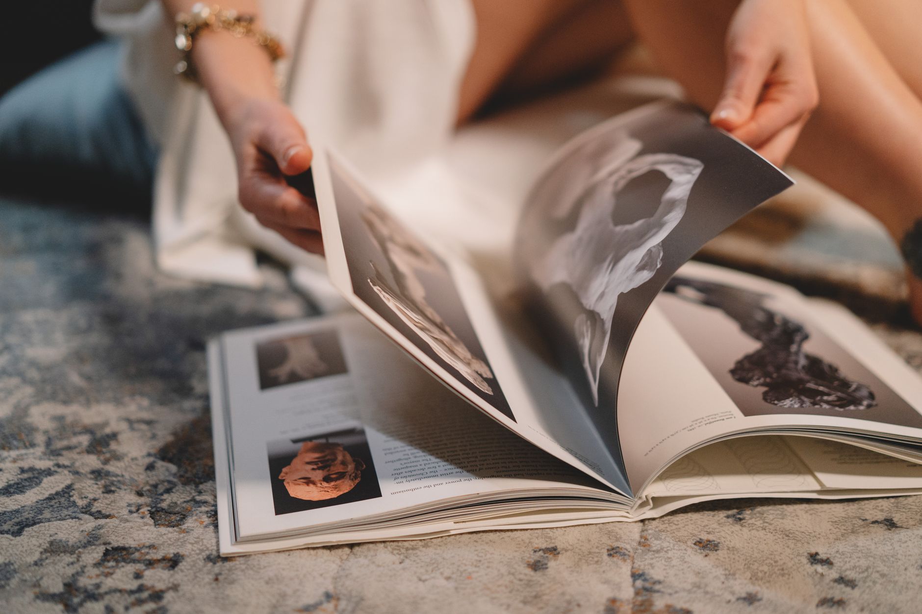 Crop unrecognizable female in white robe sitting on comfortable sofa and reading interesting book with pictures