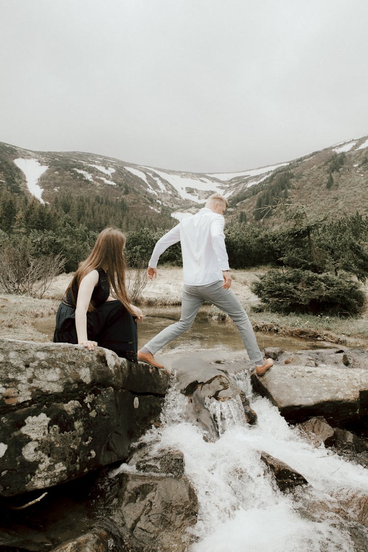 Man Wearing White Shirt And Girl In A Black Dress Jumping Over A Mountain Stream