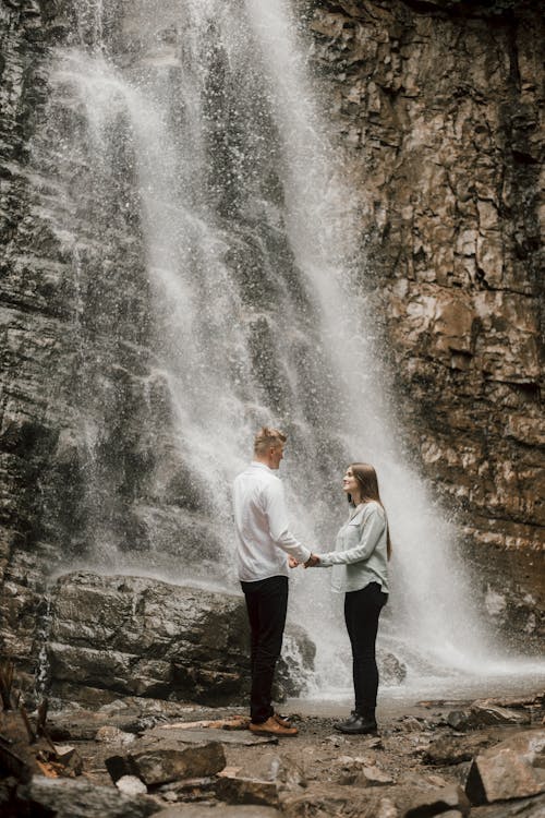 Photo of a Couple Holding Hands Beside a Waterfall