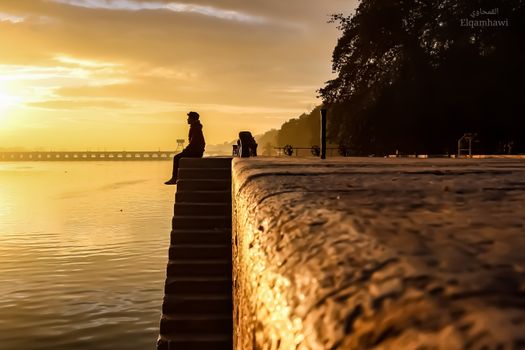 A solitary figure sits on steps by a river under a golden sunset sky, emphasizing contemplation and tranquility.