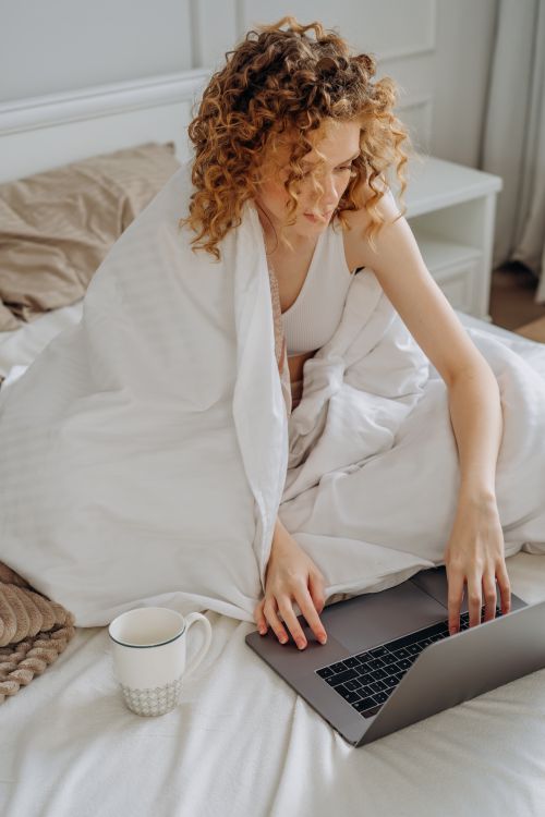Free A woman with curly hair sitting on a bed using a laptop, wrapped in a blanket, with a cup nearby. Stock Photo