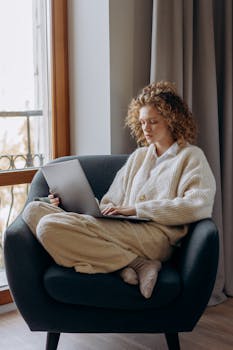 A woman in a sweater uses a laptop comfortably sitting on a couch at home.