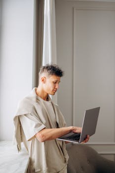 Young man standing by window using a laptop, capturing the essence of modern remote work.