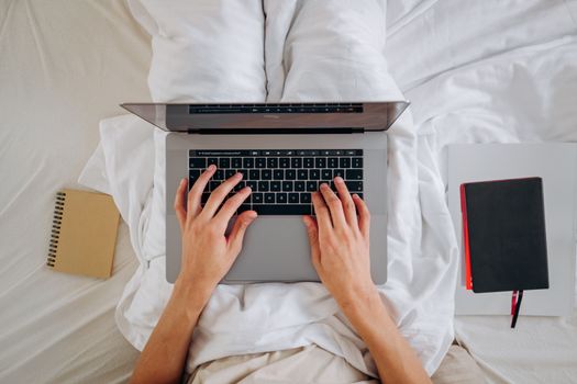 Overhead view of hands typing on a laptop while sitting on a bed with notebooks nearby, ideal for remote work themes.