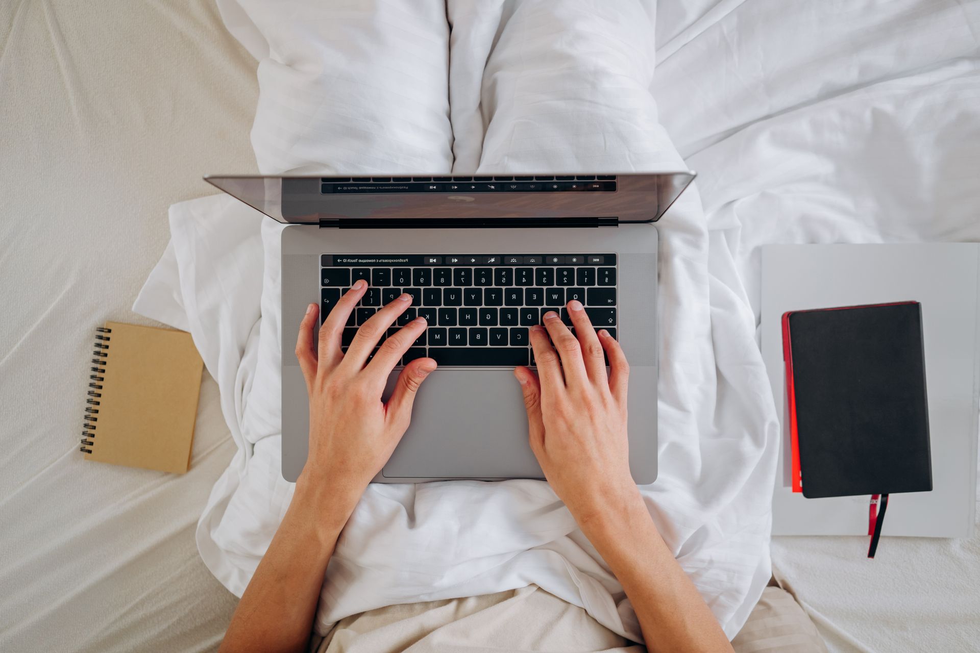 Overhead view of hands typing on a laptop while sitting on a bed with notebooks nearby, ideal for remote work themes.