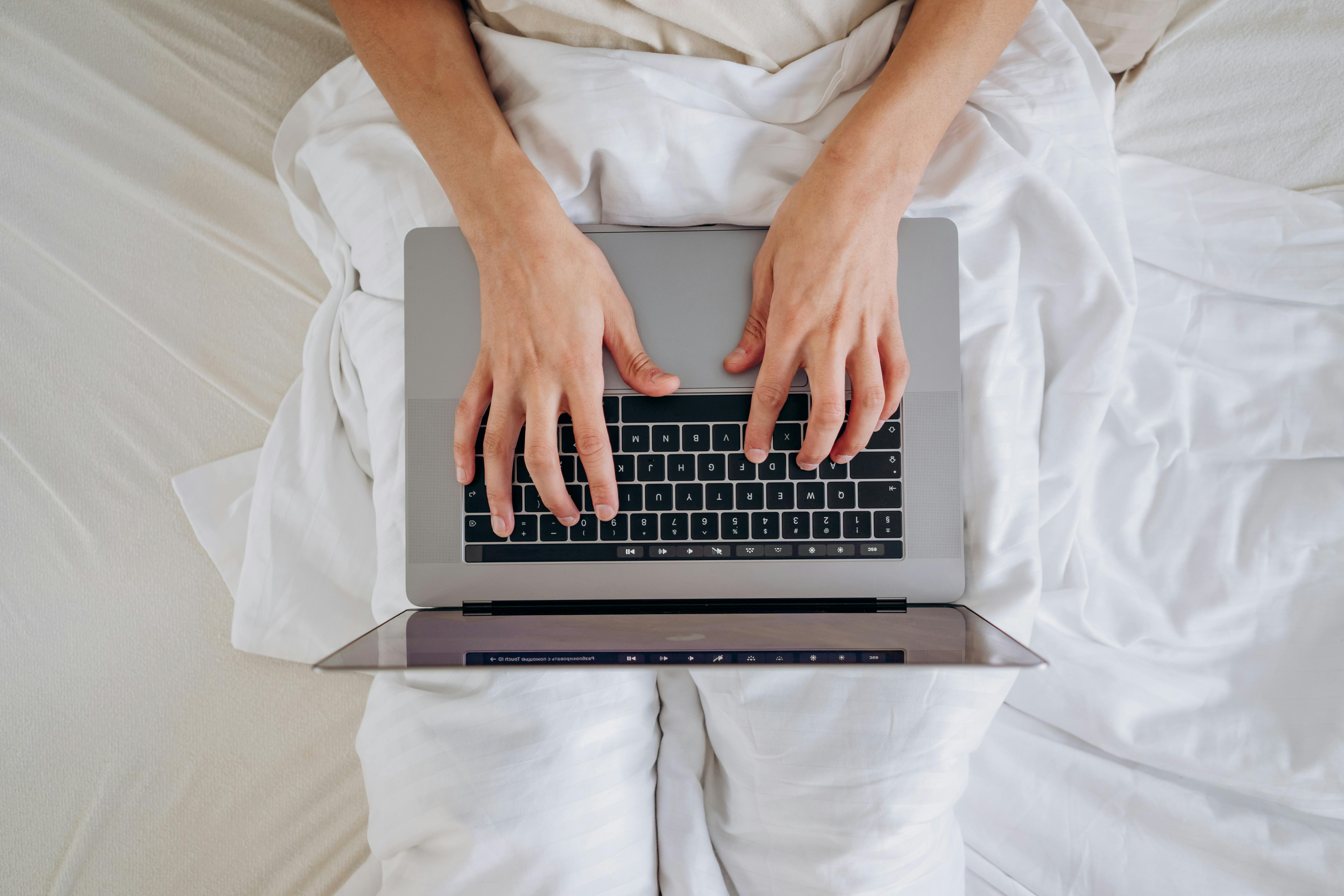 Close-up of hands typing on a laptop, resting on a lap in bed with white linens.