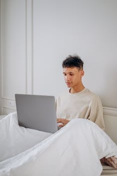 A young man sitting in bed working on a laptop indoors.