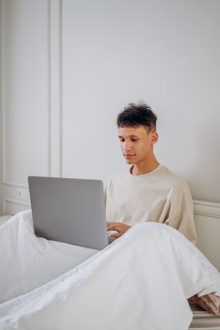 A young man sitting in bed working on a laptop indoors.