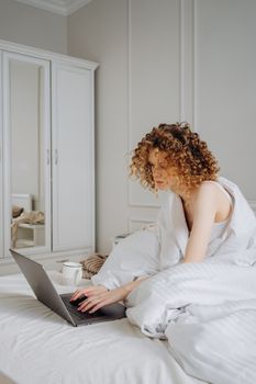 A woman typing on a laptop in a bright bedroom, wrapped in a blanket, during the daytime.