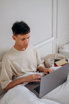 Adult man sitting in bed using a laptop in a cozy bedroom setting, embodying relaxation and technology.
