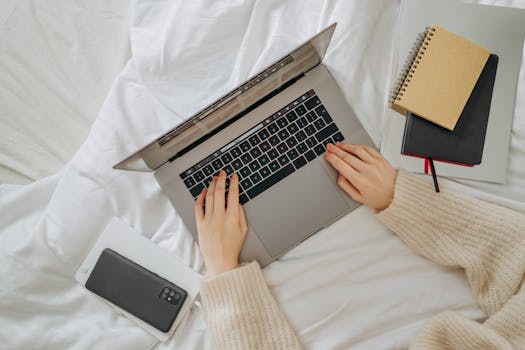 A woman using a laptop on her bed, surrounded by a smartphone and notebooks, creating a cozy workspace.
