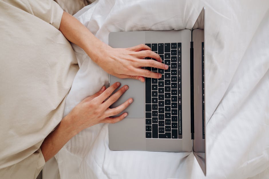 Stop Guessing Mattress Firmness—Use This Weight Chart Top view of a person typing on a laptop, seated on a bed with a white blanket.