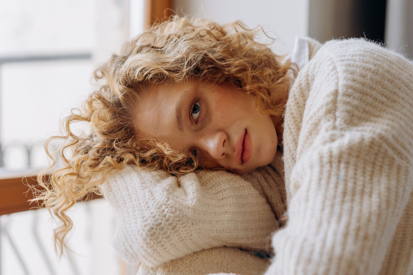 Woman with curly blonde hair resting her head on her arm, wearing a cream knit sweater by a window