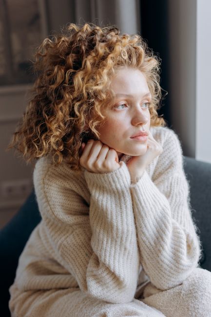 A woman with curly hair in a knit sweater looking thoughtful and relaxed indoors.