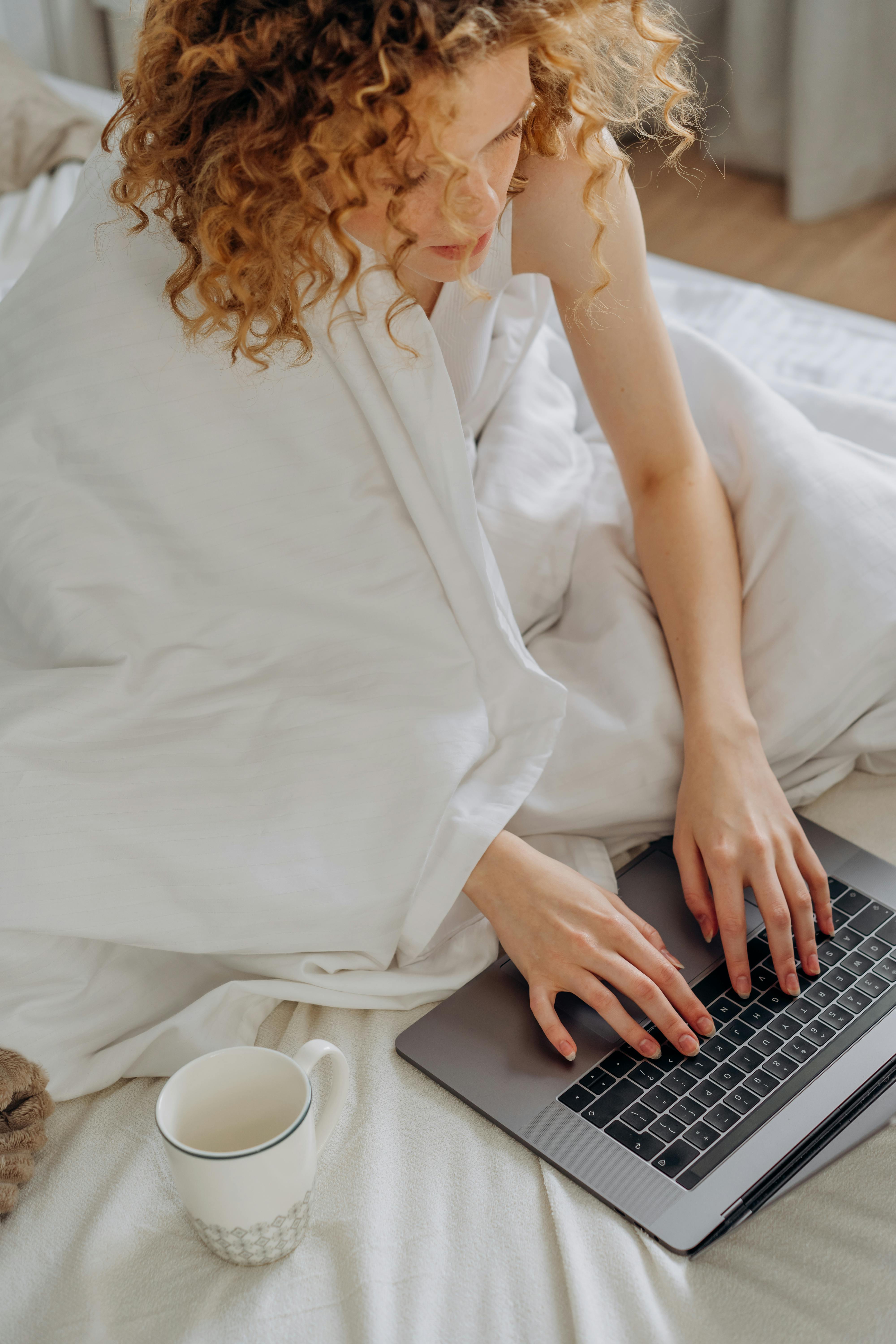 Adult woman typing on a laptop in bed with a cup of coffee nearby, creating a cozy atmosphere.