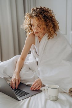 A woman comfortably working on a laptop from bed, wrapped in a blanket with a coffee cup nearby.