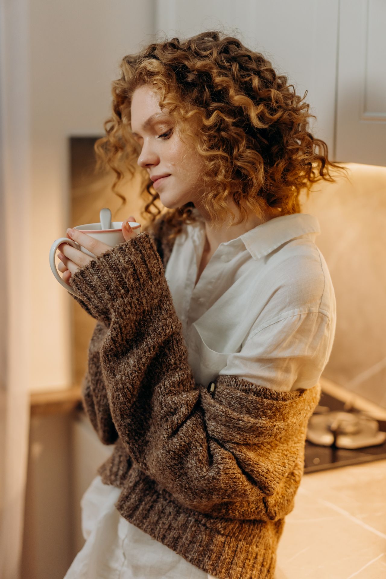 Woman layering a chunky brown knit cardigan over a white collared shirt while holding a mug