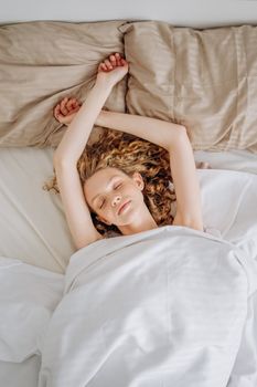A woman with curly hair sleeps peacefully with arms raised, wrapped in a white blanket.